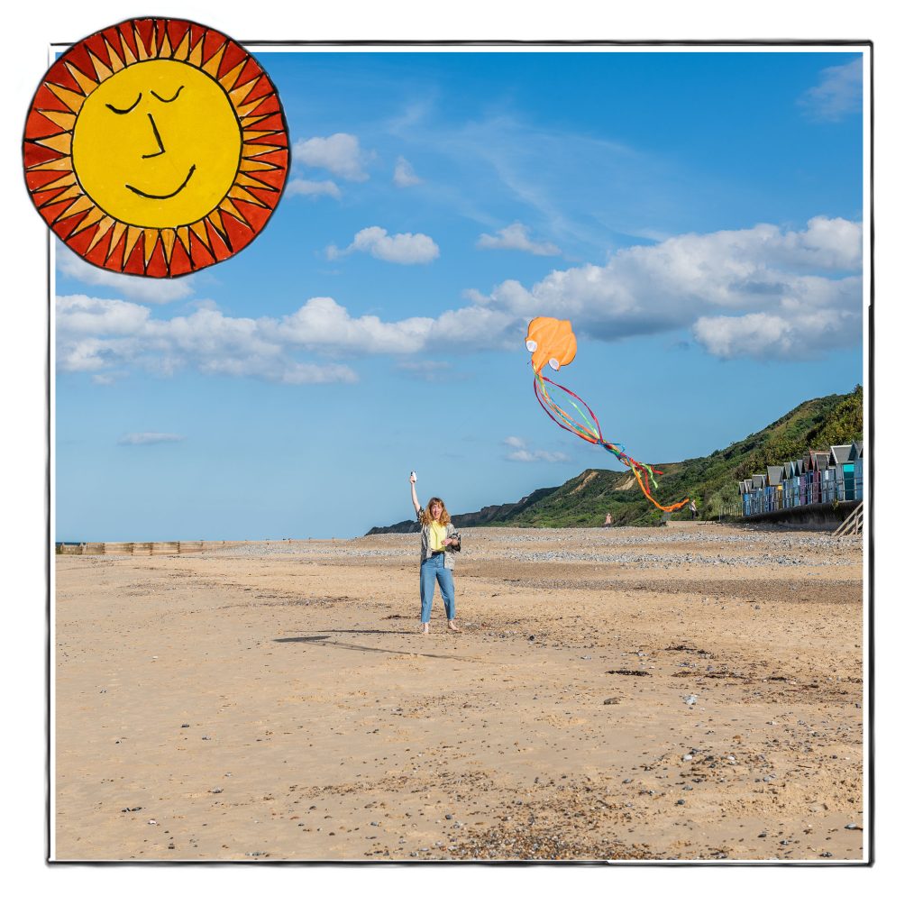 Image of a kite being flown on the beach in Norfolk, UK, framed with a sketch line and overlaid with a hand drawn image of a folk art sun