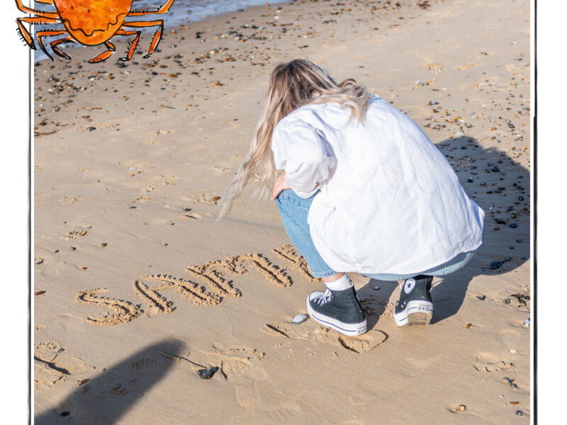 Image of a woman writing the word Samphire in the sand on the beach in Norfolk, UK, framed with a sketch line and overlaid with a hand drawn image of a crab